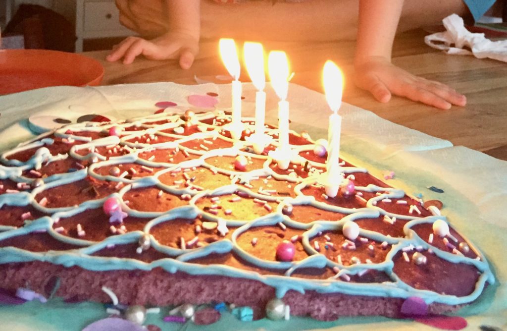 Ein aus einem lila Blechkuchen ausgeschnittene Meerjungfrauenflosse in hellblau, lila und Pink mit Zuckerperlen, Smarties und Glasur dekoriert auf einem hellblauen Untergrund mit lila, silber, blauen Konfettis dekoriert , ist ein einfacher Meerjungfrauenkuchen - der Mermaid-Cake