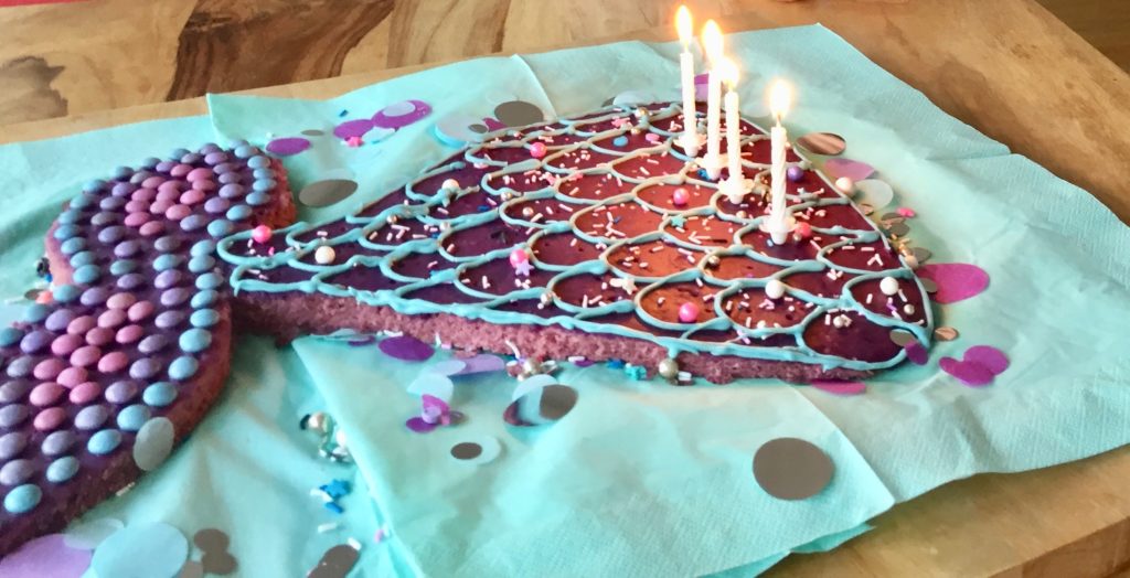 Ein aus einem lila Blechkuchen ausgeschnittene Meerjungfrauenflosse in hellblau, lila und Pink mit Zuckerperlen, Smarties und Glasur dekoriert auf einem hellblauen Untergrund mit lila, silber, blauen Konfettis dekoriert - der Mermaid-Cake