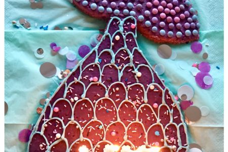 Ein aus einem lila Blechkuchen ausgeschnittene Meerjungfrauenflosse in hellblau, lila und Pink mit Zuckerperlen, Smarties und Glasur dekoriert auf einem hellblauen Untergrund mit lila, silber, blauen Konfettis dekoriert