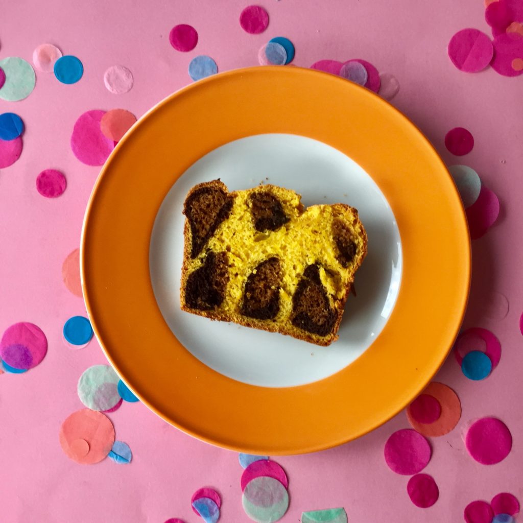 Auf einem weißen Teller mit dickem orangenen Rand befindet sich eine Scheibe Kuchen mit braunen Leopardenmuster. Der Teller befindet sich auf einem rosa Untergrund mit bunten Konfettis dekoriert.