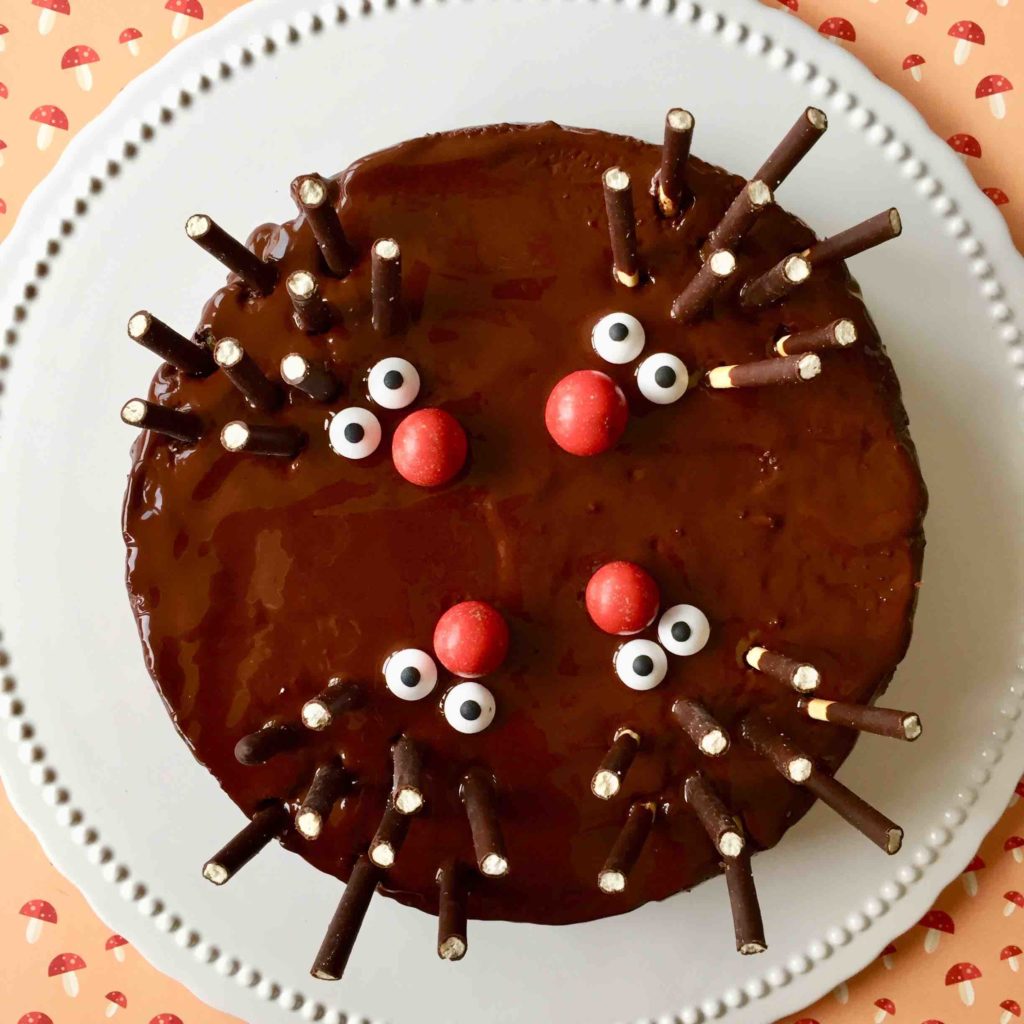 Ein Schoko-Nuss-Kuchen mit vier kleinen Igeln als Igelkuchen  gestaltet aus einer roten Nase aus M und Ms, Zuckeraugen und Micadostäbchen als Stacheln auf einer weißen Kuchenplatte auf einem Papieruntergrund mit aufgedruckten Pilzen.