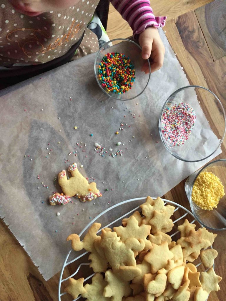 Ein Kind bestreuselt die selbstgebackenen Plätzchen mit Zuckerstreuseln. Wie das Dekorieren und Plätzchen backen mit Kindern in der Weihnachtszeit zum Erfolg wird, zeigen dir diese Backtipps.